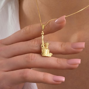May include: A gold-colored pendant necklace featuring a detailed miniature of the Statue of Liberty. The pendant is suspended from a delicate chain. The necklace is held up by a person's hand.