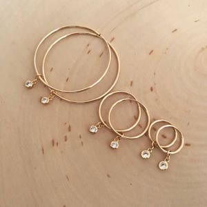 May include: Set of four gold hoop earrings with a small clear crystal dangling from each hoop. The hoops are different sizes, with the largest hoop being the topmost hoop.