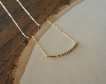 Puede incluir: Un collar de oro con un simple colgante de barra curva. El collar está sobre una superficie de madera.