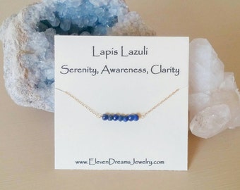 May include: A gold chain necklace with a bar of lapis lazuli beads. The card reads "Lapis Lazuli Serenity, Awareness, Clarity" and "www.ElevenDreamsJewelry.com".