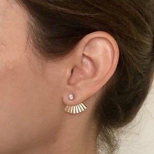 May include: Gold ear cuff earrings with a fan-shaped design and a small clear crystal stud. The earrings are worn in the upper lobe of the ear.