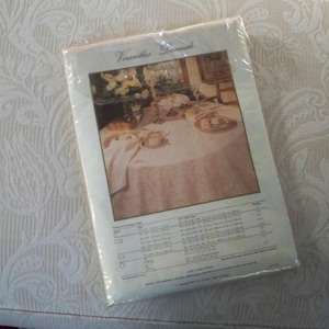 May include: A packaged tablecloth with the words "Versailles Damask" printed on the front. The image on the package shows a round table set with a tablecloth, dishes, and flowers. The tablecloth is a light pink color.