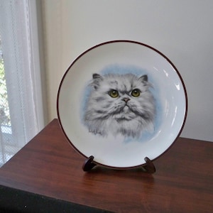 May include: A decorative plate featuring a detailed portrait of a Persian cat with gray fur and yellow eyes. The plate has a white background with a blue oval around the cat's face and a dark red rim. It is displayed on a wooden stand.