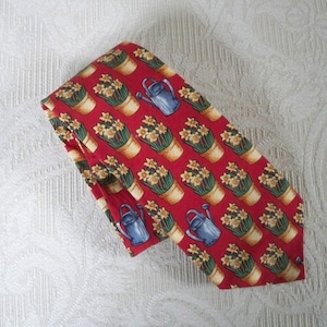 May include: A red necktie with a repeating pattern of potted daffodils and blue watering cans. The daffodils are yellow and green, and the pots are tan. The tie is made of a smooth fabric.