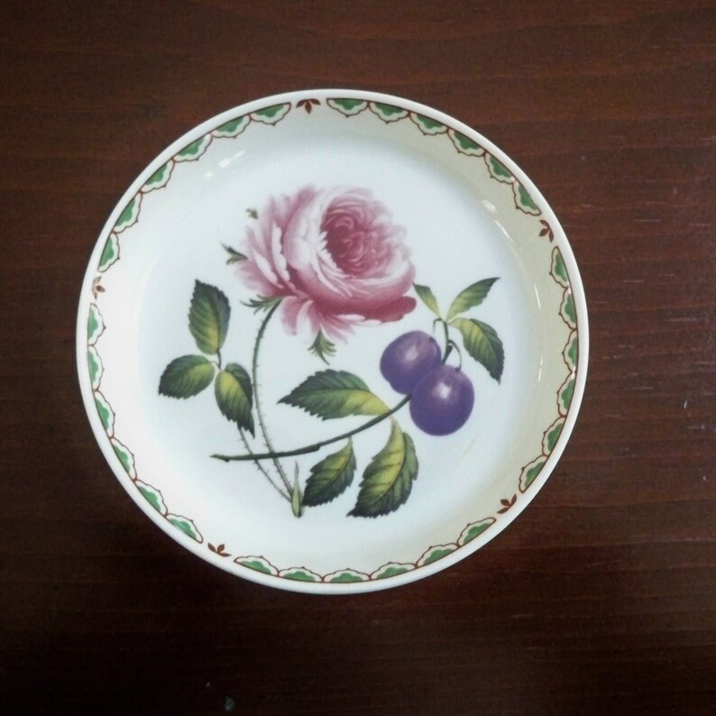 May include: A white ceramic plate with a green and brown border. The plate features a pink rose with green leaves and two purple plums.