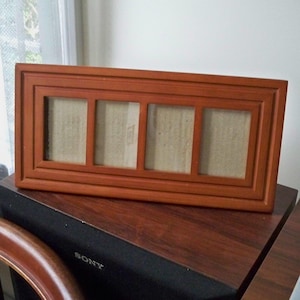 May include: A rectangular wooden picture frame with a warm brown finish, featuring four vertical openings arranged side-by-side. The frame has a stepped, layered border design and contains plain, light-colored backing inserts in each section.