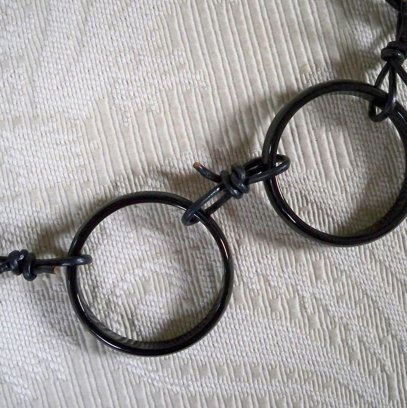 May include: A black leather necklace with two black, round, polished metal rings connected by the leather cord. The rings are interlocked.