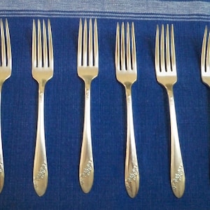 May include: Six silver forks with ornate designs on a blue and white striped tablecloth.