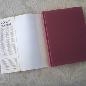 May include: An open book titled "Vogue Sewing" with a burgundy cover. The book is on a patterned surface. The left page is open, revealing white pages. The book's spine is visible, and the cover is a solid burgundy color.