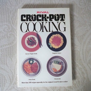 May include: A cookbook cover with the title "RIVAL CROCK-POT COOKING" in black and white text. The cover features four photos of food in ceramic dishes: Savory Pepper Steak, Spaghetti Sauce, Swiss Steak, and Cheesecake. The text "More than 300 recipes especially for the original Crock-Pot slow cooker" is at the bottom of the cover.