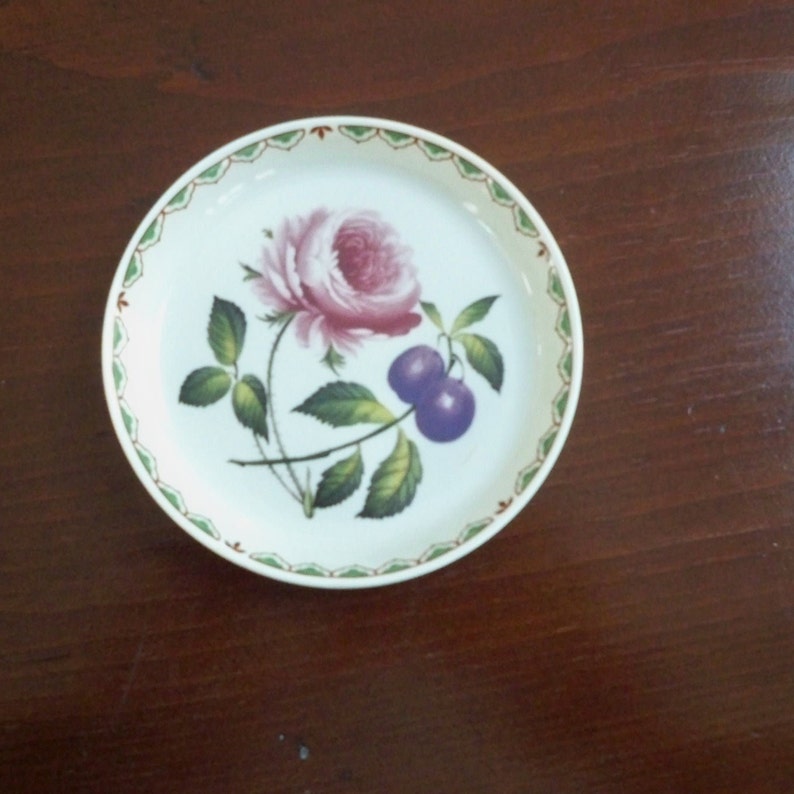 May include: A white ceramic coaster with a green and red border. The coaster features a pink rose with green leaves and two purple plums.