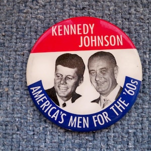 May include: A vintage campaign button featuring portraits of John F. Kennedy and Lyndon B. Johnson. The button has a red and white top with the text "KENNEDY JOHNSON." The bottom arc reads "AMERICA'S MEN FOR THE '60s" in blue.