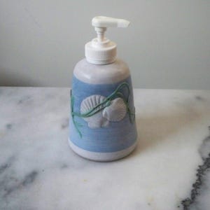 May include: A ceramic soap dispenser with a white pump top. The body is light blue with white seashell and seaweed accents. The dispenser is cylindrical with a slightly flared base.