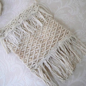May include: Cream-colored macrame bag with a geometric diamond pattern and fringe detailing. The bag is rectangular with a woven design and long fringe along the top and bottom edges. The bag is made of a natural fiber.