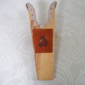May include: Wooden boot jack with a brown leather patch featuring a horse head design. The boot jack has a light brown wood finish and is padded with beige fabric.