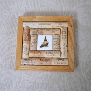 May include: Square wooden framed artwork made from wine corks. The corks surround a small square tile with a wine bottle, grapes, and a glass. The frame is light brown, and the corks have various winery names printed on them.