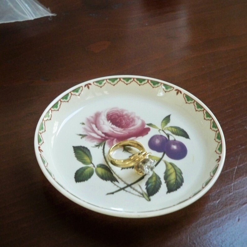 May include: A white ceramic trinket dish with a green and brown floral design. The dish features a pink rose, green leaves, and two purple plums. A gold ring with a diamond is sitting on the dish.