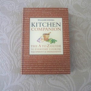 May include: A hardcover cookbook titled "Kitchen Companion" by Williams-Sonoma. The book features a white cover with the title and a graphic of a mortar and pestle, garlic, and herbs. The book is an A to Z guide to everyday cooking equipment and ingredients.