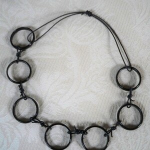 May include: A black necklace made of metal rings connected by black string. The necklace has a simple design and is made of multiple rings.