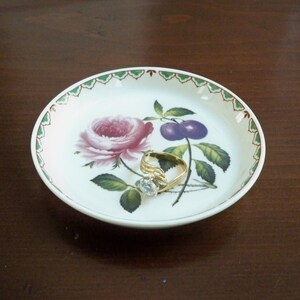 May include: A white ceramic trinket dish with a green and red border. The dish features a floral design with a pink rose, green leaves, and purple plums. A gold ring with a clear stone is resting on the dish.