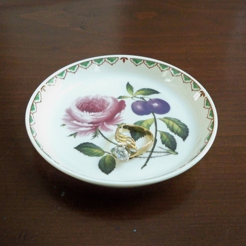 May include: A white ceramic trinket dish with a green and red border. The dish features a floral design with a pink rose, green leaves, and purple plums. A gold ring with a clear stone is resting on the dish.