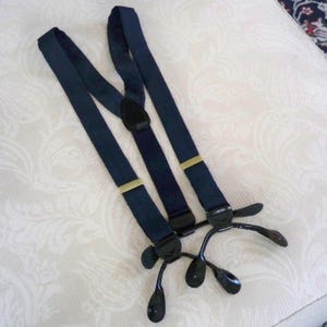 May include: A pair of navy blue suspenders featuring adjustable gold-tone metal sliders and black plastic clips at the ends. The Y-back design includes a central black leather-like patch where the elastic straps meet, providing a secure hold.