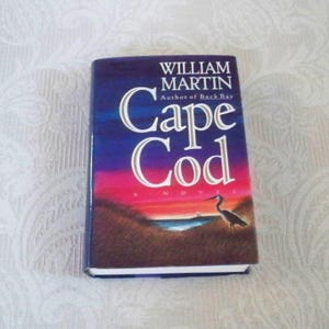May include: A hardcover book titled "Cape Cod" by William Martin. The cover features a colorful sunset over a coastal landscape with a bird silhouette. The book's title is in large white letters.