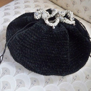 May include: A black, round drawstring bag with a textured surface. The bag is adorned with a silver-colored, decorative clasp embellished with small, gray beads. The bag's drawstring is visible, and the background is a patterned fabric.