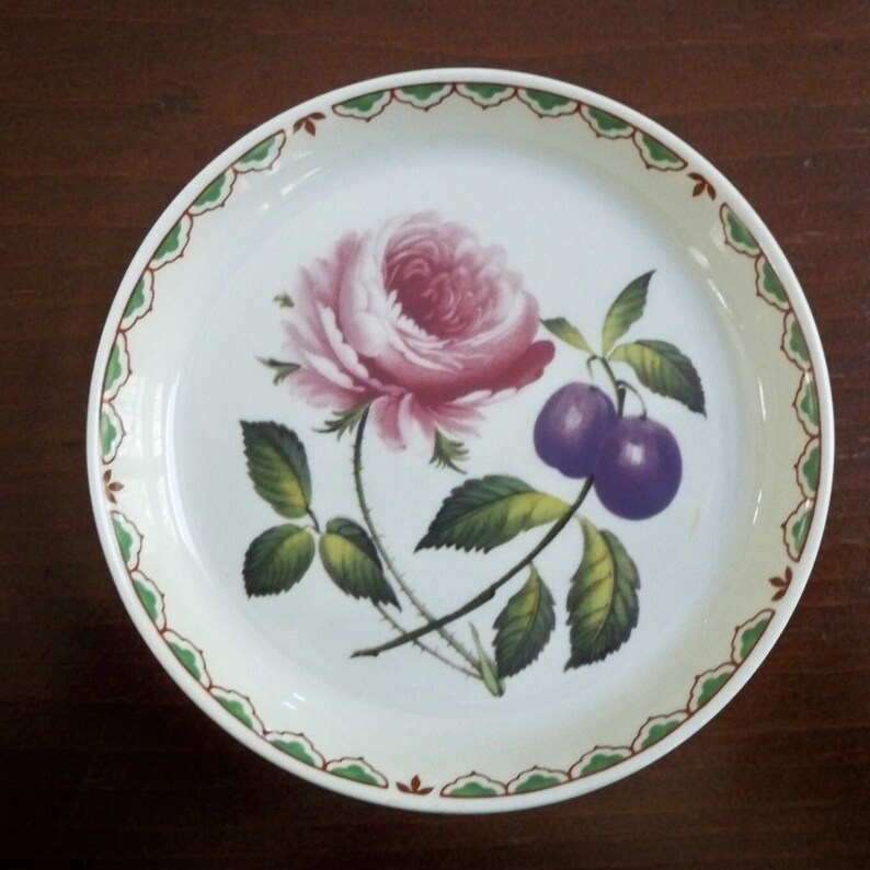 May include: A white ceramic plate with a green and brown border. The plate features a hand-painted design of a pink rose and two purple plums.