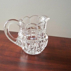 May include: A clear glass creamer with a handle and a decorative, textured pattern. The creamer has a scalloped rim and sits on a dark wooden surface. The creamer is designed for serving cream or milk.
