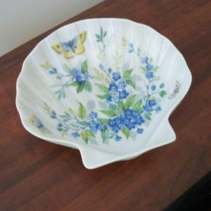 May include: White, shell-shaped decorative dish with a floral and butterfly design. The dish features blue flowers, green leaves, and yellow and blue butterflies. The dish has a scalloped edge.