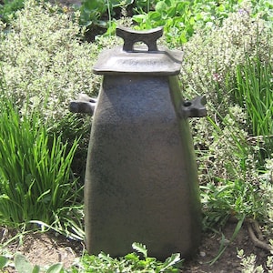 May include: A black ceramic urn with a lid and a textured surface. The urn is sitting on a bed of green plants.