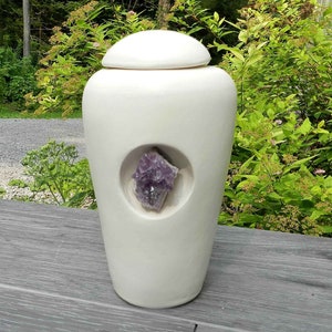 May include: White ceramic urn with a round lid and a cutout on the front revealing a purple amethyst crystal.