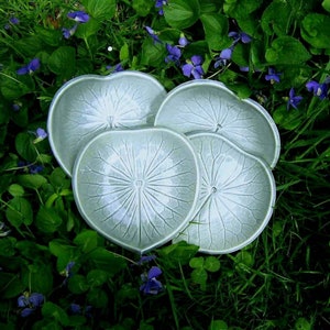 May include: Four ceramic plates in a light green colour with a leaf design. The plates are arranged in a circle on a bed of green grass and purple flowers.