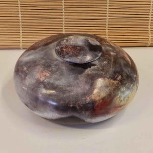 May include: A round, low-profile ceramic vessel with a matching lid, featuring a mottled, smoke-fired finish in shades of charcoal, deep brown, and hints of burnt orange. The surface has a smooth, polished appearance with organic, cloud-like patterns.
