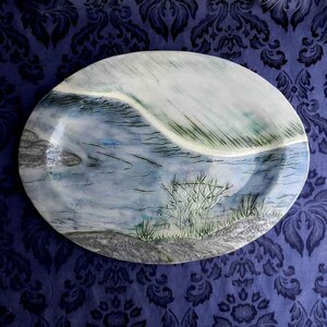 May include: A ceramic oval platter with a blue and white landscape design. The platter features a stylised depiction of a river with a white shoreline and green foliage.