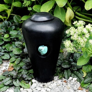 May include: A black urn with a turquoise stone inlay. The urn has a rounded top and a smooth, matte finish. It is surrounded by green foliage and small white flowers, creating a natural setting.