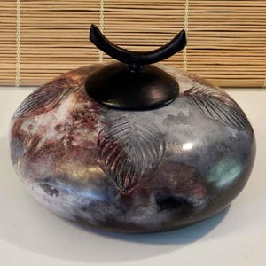 May include: A round, low-profile ceramic vessel with a smooth, mottled surface featuring shades of black, gray, and deep red. The body is decorated with carved leaf patterns and topped with a black, crescent-shaped lid handle.