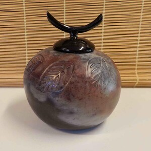 May include: A round ceramic jar with a mottled brown and grey finish featuring embossed leaf patterns on the surface. The jar includes a removable lid topped with a smooth, glossy black handle shaped like a crescent moon or a stylized horn.