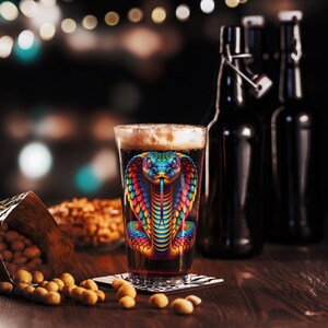 Könnte beinhalten: Ein Glas dunkles Bier mit einem bunten Kobra-Schlangen-Design an der Seite. Das Glas steht auf einem Holztisch mit Erdnüssen, die um ihn herum verstreut sind.