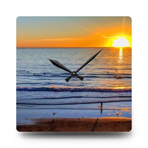 Könnte beinhalten: Eine quadratische Uhr mit einem Sonnenuntergang über dem Meer als Hintergrund. Das Zifferblatt der Uhr ist weiß mit schwarzen Zeigern. Das Meer ist blau und der Himmel ist orange und gelb. Es sind Menschen im Wasser und am Strand zu sehen.