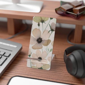 May include: A rectangular phone stand featuring a light cream background with a repeating pattern of soft-toned, watercolor-style flowers in shades of pale yellow, muted peach, and light green, complete with dark brown centers and thin stems.