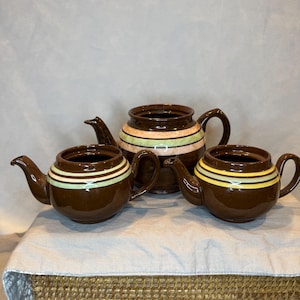 Puede incluir: Tres teteras de cerámica marrón con diseños de rayas coloridas. Las teteras están dispuestas sobre una superficie tejida.