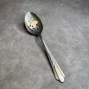 May include: A silver slotted spoon with a floral design on the bowl. The handle has a decorative pattern. The spoon is designed for straining liquids or serving. The spoon is made of metal and is likely vintage.