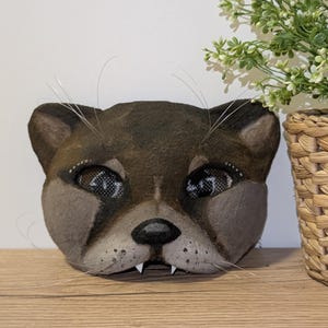 May include: A handcrafted, brown cat mask with detailed facial features, including whiskers, and mesh eye coverings. The mask is displayed on a wooden surface, with a potted plant in the background.