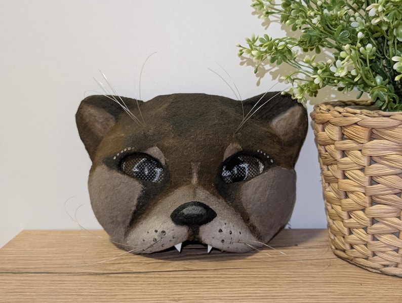 May include: A handcrafted, brown cat mask with detailed facial features, including whiskers, and mesh eye coverings. The mask is displayed on a wooden surface, with a potted plant in the background.