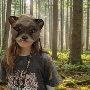 May include: A brown cat mask with whiskers and a pink mouth. The mask is worn over the face. The person is wearing a gray t-shirt with a graphic design and a beaded necklace. The background is a forest with tall trees.