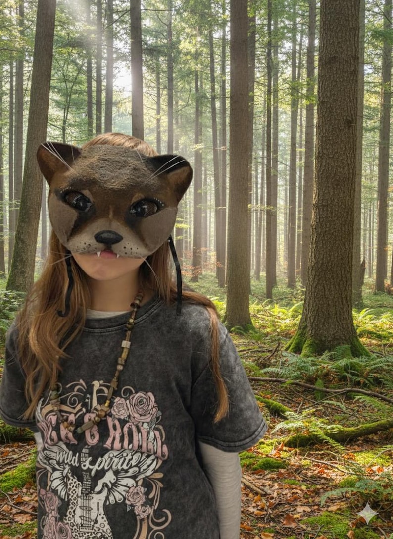 May include: A brown cat mask with whiskers and a pink mouth. The mask is worn over the face. The person is wearing a gray t-shirt with a graphic design and a beaded necklace. The background is a forest with tall trees.
