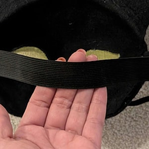 May include: Black felt mask with an elastic band. The mask has yellow mesh eye coverings. The elastic band is black and stretches across the back of the mask. The mask is likely for a costume or play.