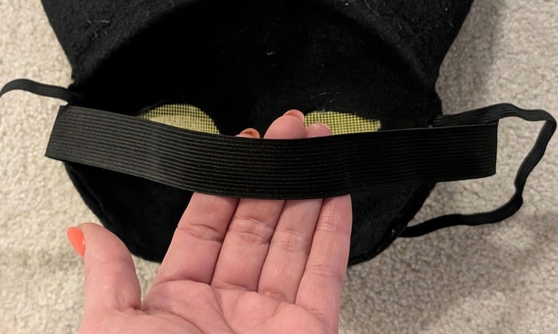 May include: Black felt mask with an elastic band. The mask has yellow mesh eye coverings. The elastic band is black and stretches across the back of the mask. The mask is likely for a costume or play.